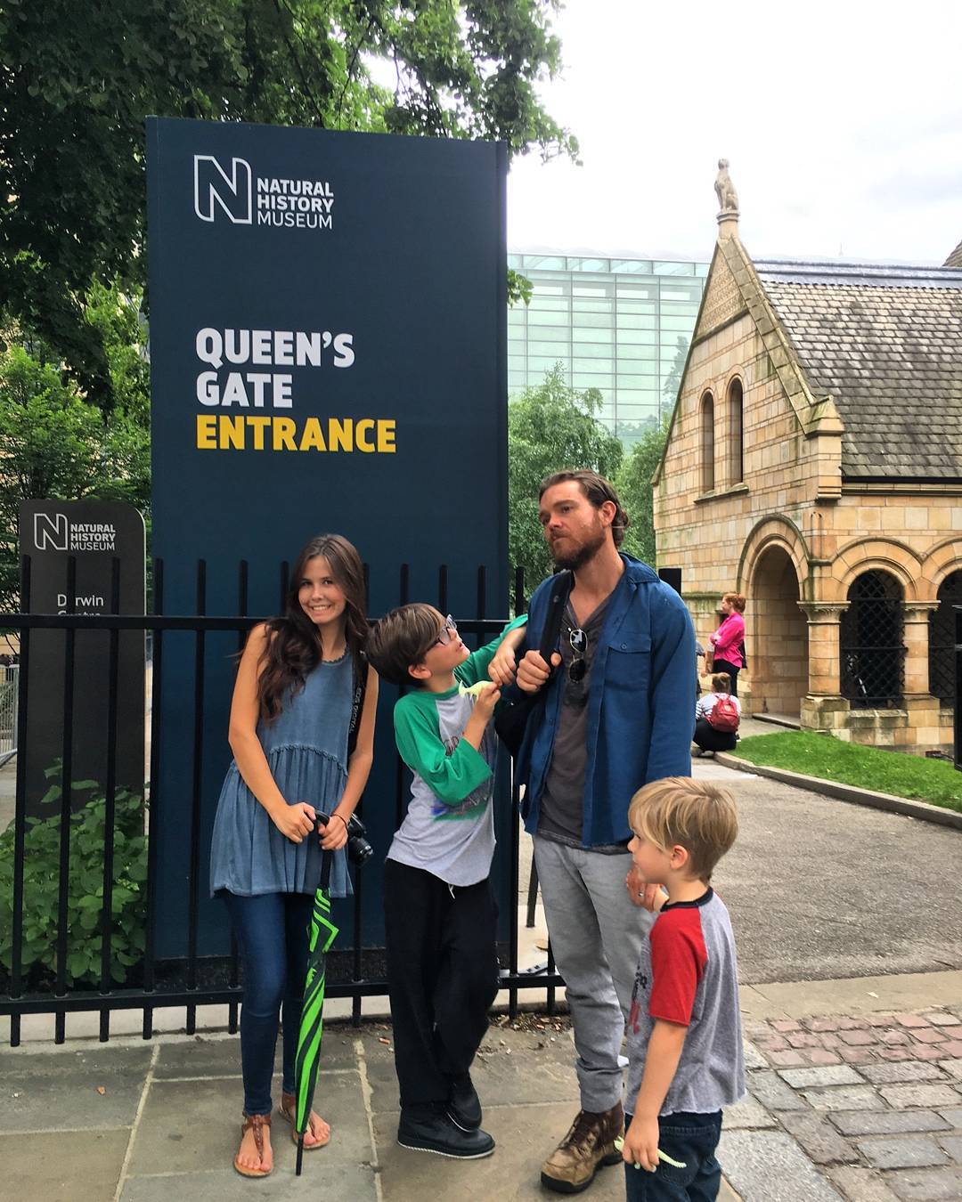 Clayne Crawford in a dark blue jacket and light blue jeans posing with his children at Queen's Gate Entrance.